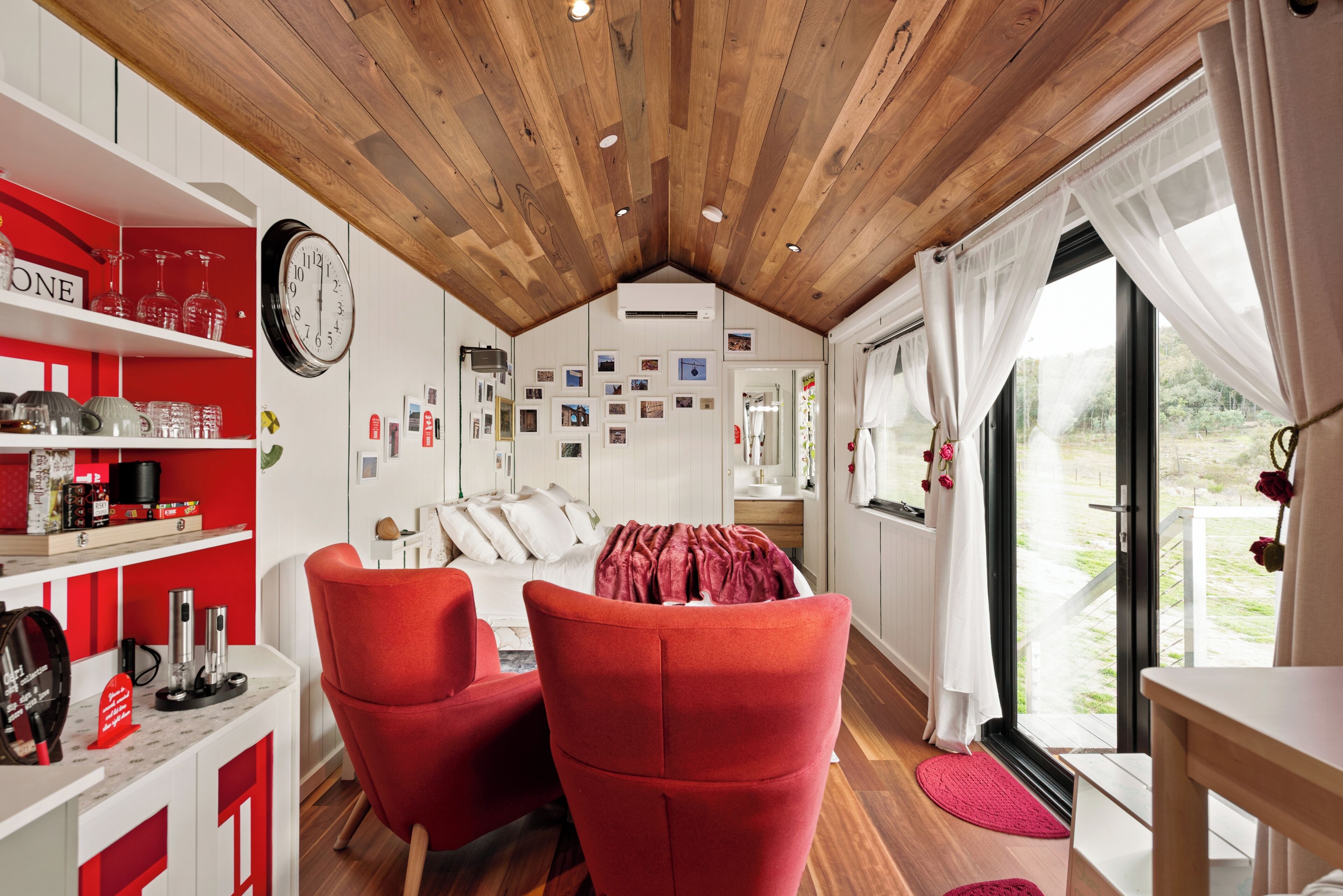 A cozy room with a wooden ceiling, a bed with white bedding, red chairs, a red shelf with various items, and a wall with pictures.