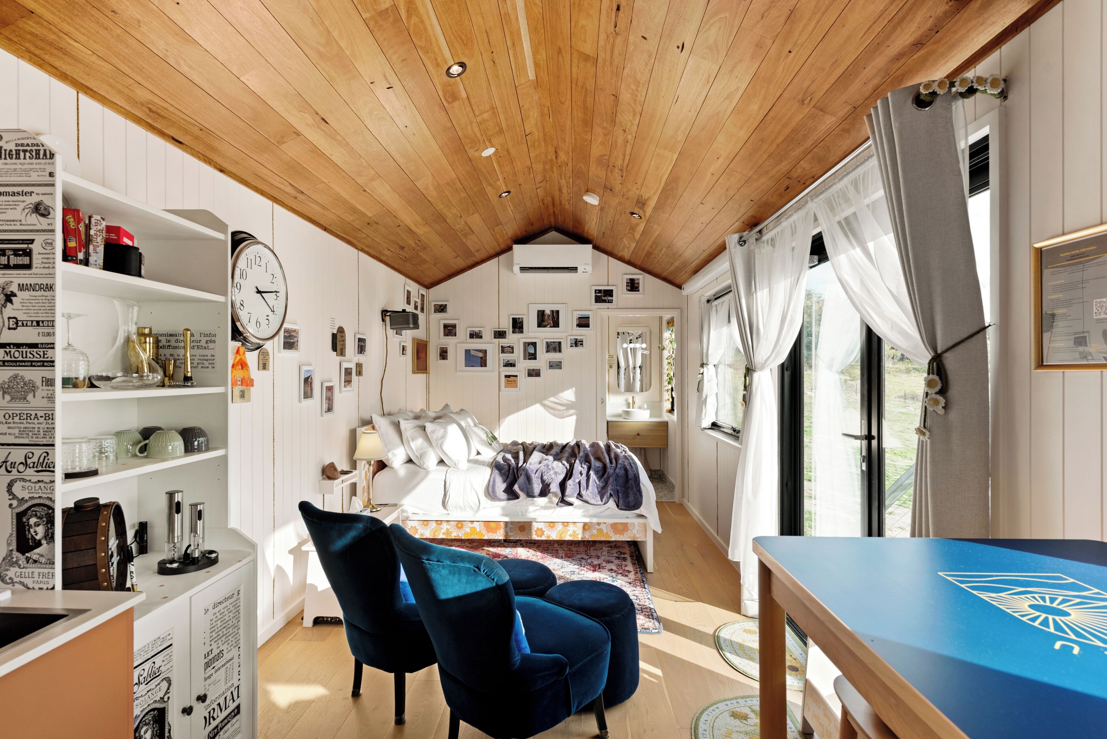 A cozy room with a wooden ceiling, a bed with a floral bedspread, a blue chair, and a wall adorned with framed pictures.