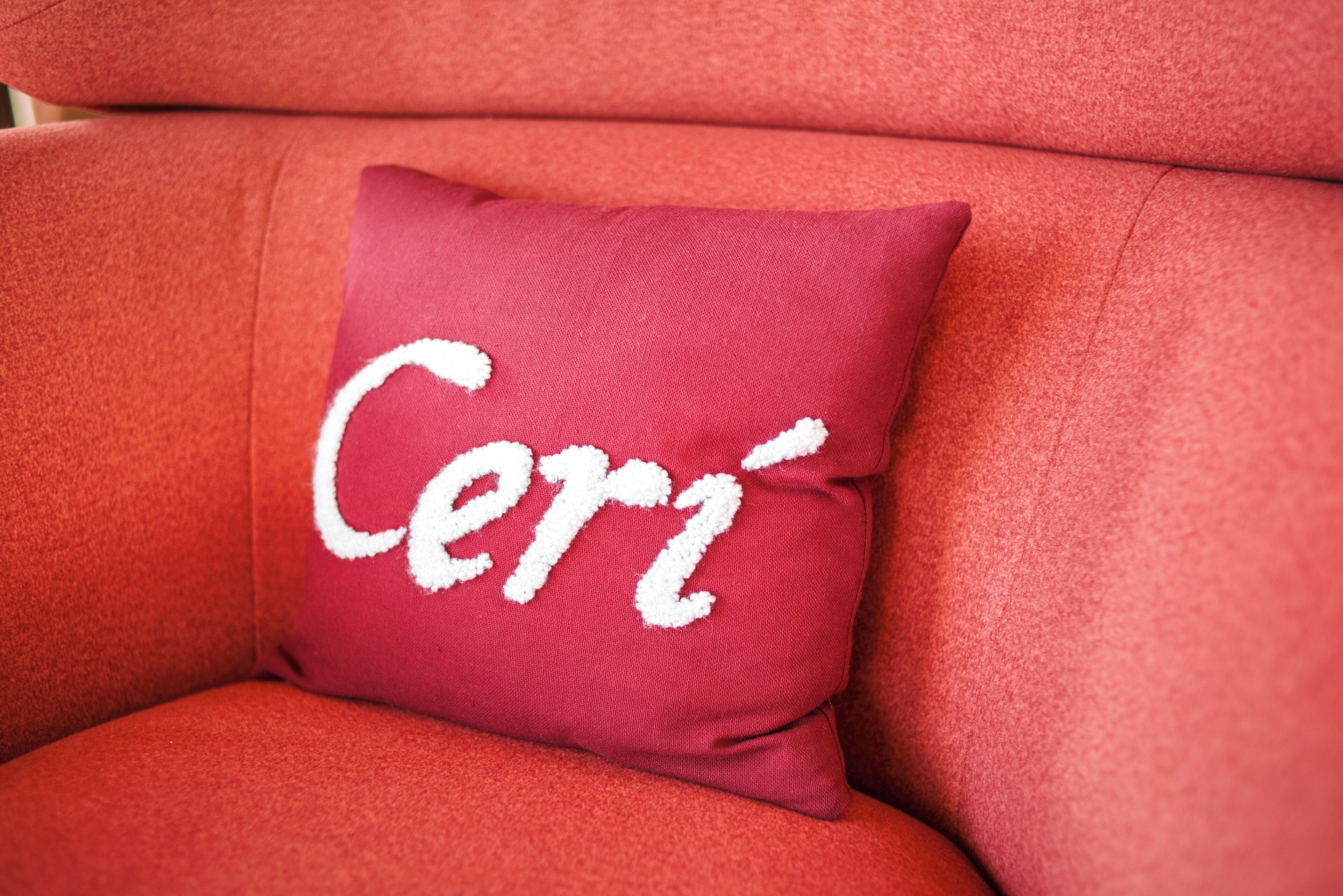 A close-up of a red upholstered chair with a white embroidered pillow that reads 'Ceri'.