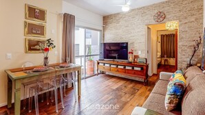 Una sala de estar con televisión, mesa de comedor de madera, sillas transparentes y una pared de ladrillo.
