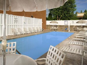 A swimming pool with white chairs and umbrellas, a lifeguard warning sign, and a brick wall in the background.
