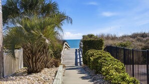 Un camí de fusta que condueix a una platja, amb una tanca d'un costat i una palmera de l'altre.