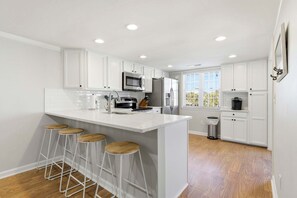A modern kitchen with white cabinetry, a central island with stools, stainless steel appliances, and a window with blinds. - Pal's Palmetto Place - 3BR Bungalow w/ Ocean Views (North Myrtle Beach)
