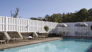 Une piscine entourée d’une clôture blanche et d’arbres, avec des chaises longues et des parasols.