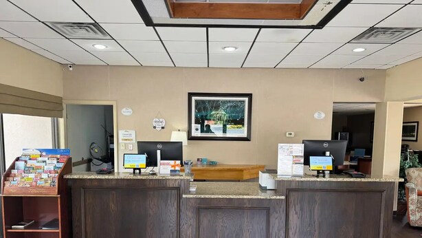 A reception area with a wooden reception desk, two computer monitors, a framed picture on the wall, and a small bookshelf with magazines. - Affordable Stay Near Liberty University with Free Breakfast & Outdoor Pool (Lynchburg)