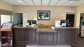 A reception area with a wooden reception desk, two computer monitors, a framed picture on the wall, and a small bookshelf with magazines.