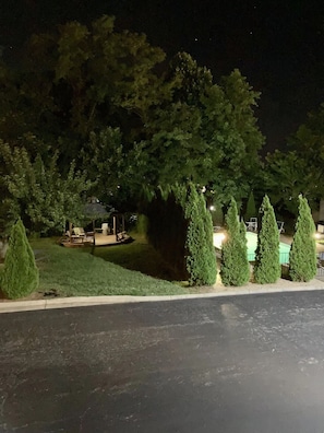 Vista nocturna de un jardín con piscina, rodeado de árboles y un patio con mobiliario exterior.