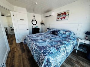 A bedroom with a bed, a bedside table, a nightstand, a wall-mounted air conditioner, and a door leading to another room.