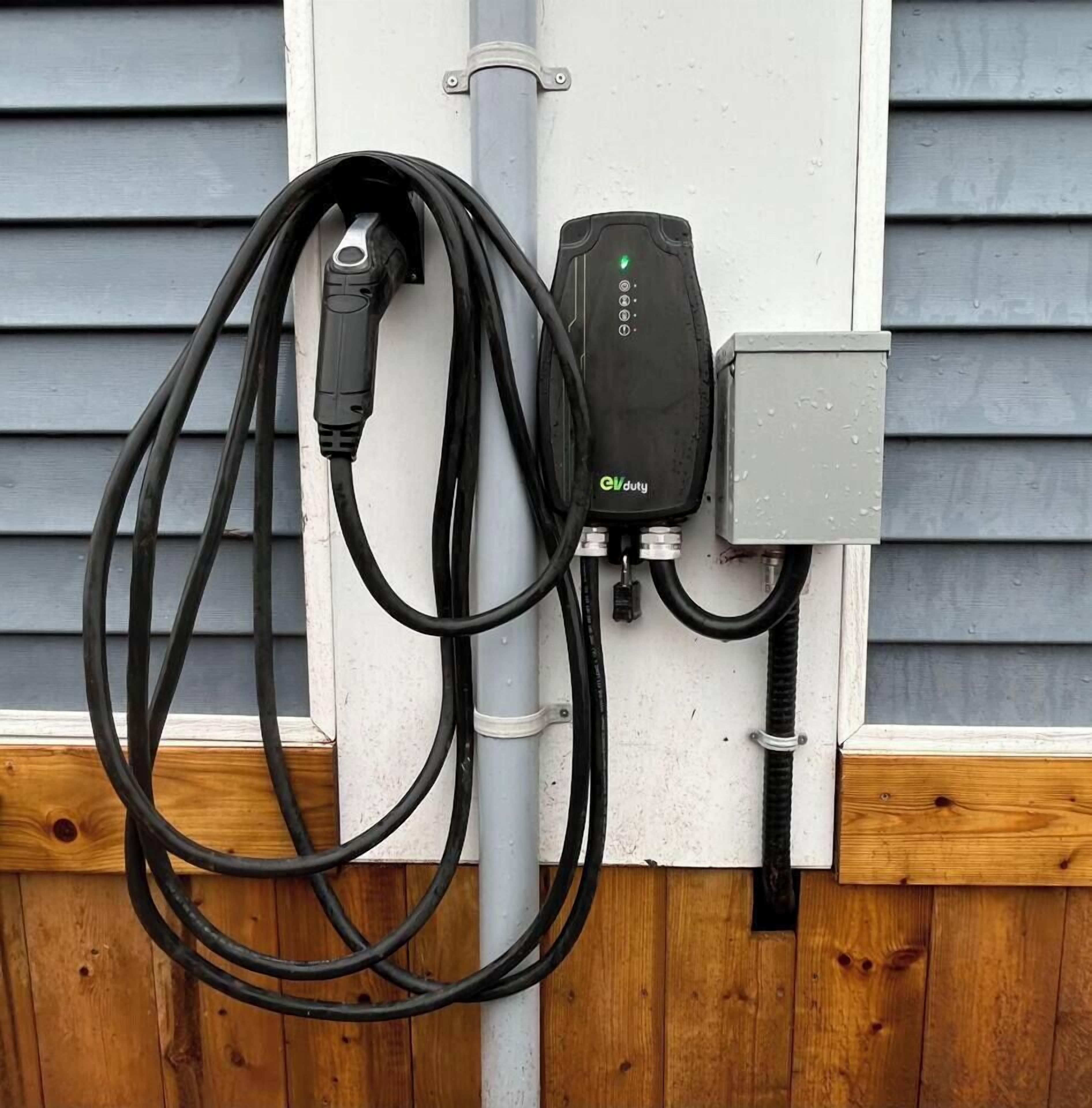 An electric vehicle charging station with a cable connected to a charging port, mounted on a wall with a wooden fence below.