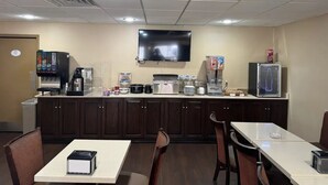 Una habitación pequeña y bien iluminada con kitchenette, un televisor de pantalla plana montado en la pared y algunas mesas y sillas.