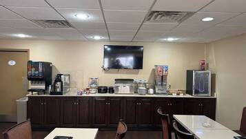 A small, well-lit room with a kitchenette, a flat-screen TV mounted on the wall, and a few tables and chairs.