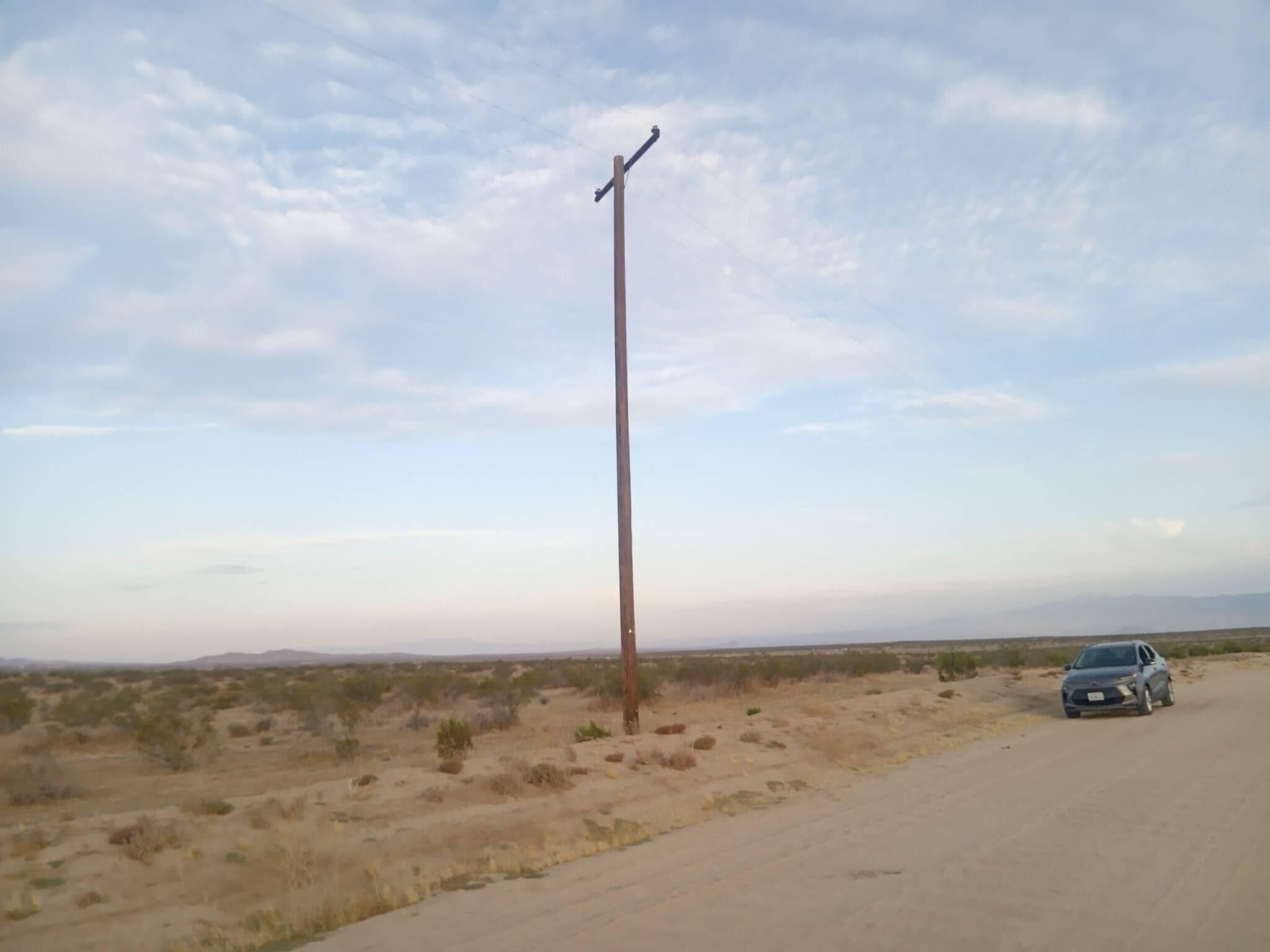 A desert landscape with sparse vegetation, a single utility pole, and a parked car on a dirt road.