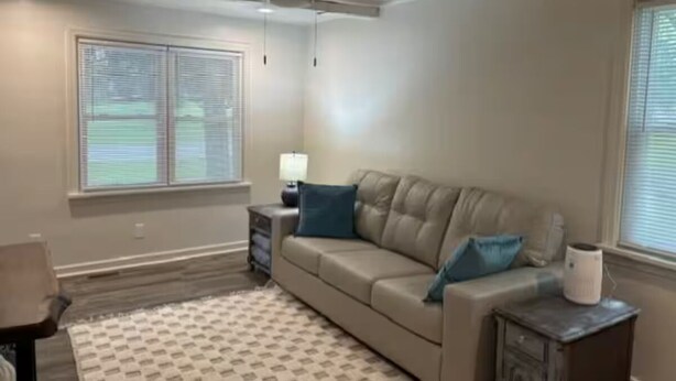 Una sala de estar con sofá color beige, alfombra estampada, ventilador de techo y una ventana con persianas.