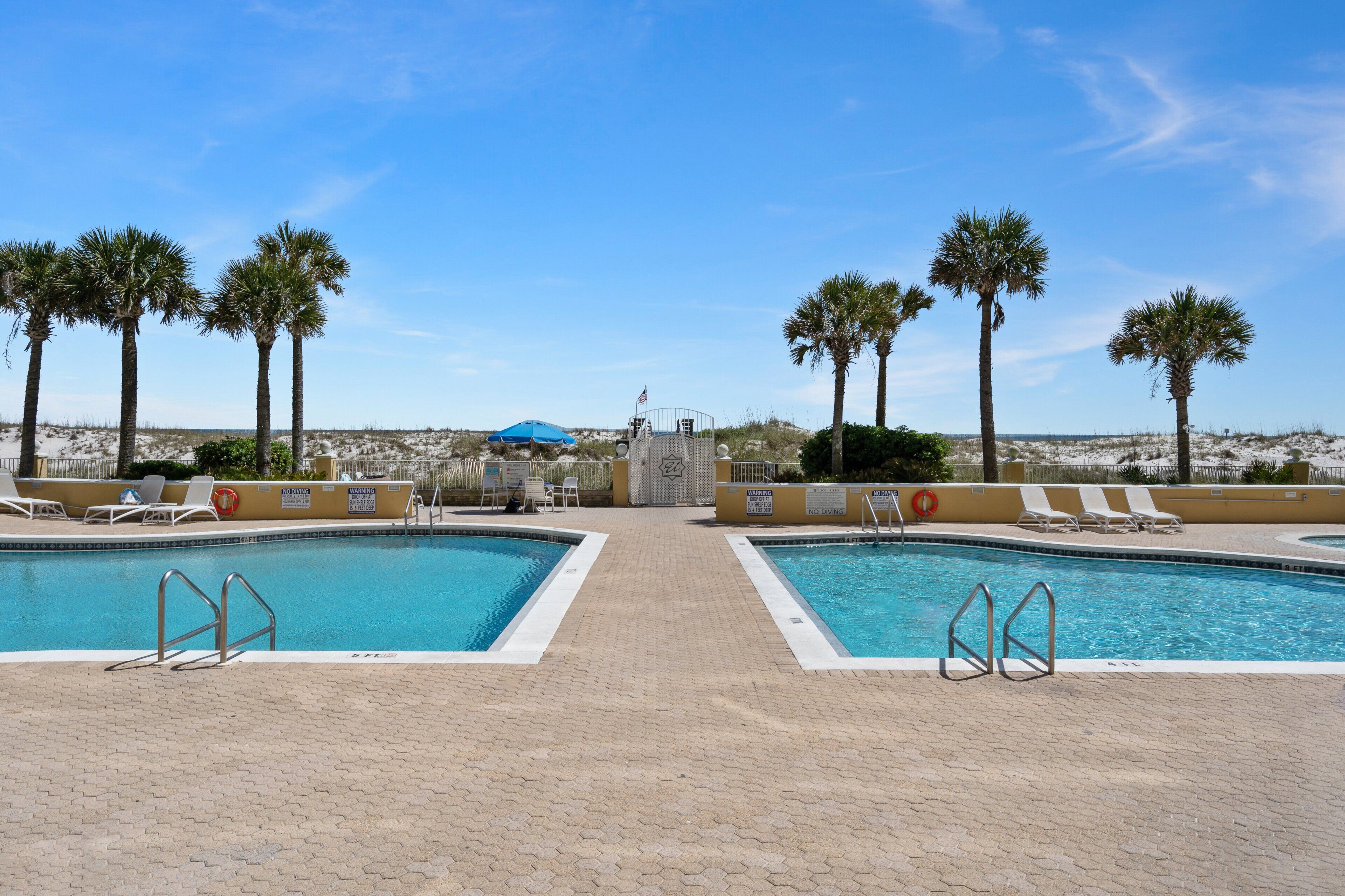 A pool area with two separate pools, lounge chairs, palm trees, and a distant view of a beach.
