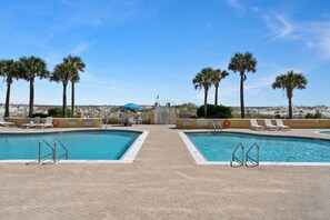 A pool area with two separate pools, lounge chairs, palm trees, and a distant view of a beach. - Emerald Isle 504 | Pool, Gym | Seasonal Beach Svc. | My Beach Getaways (Pensacola Beach)