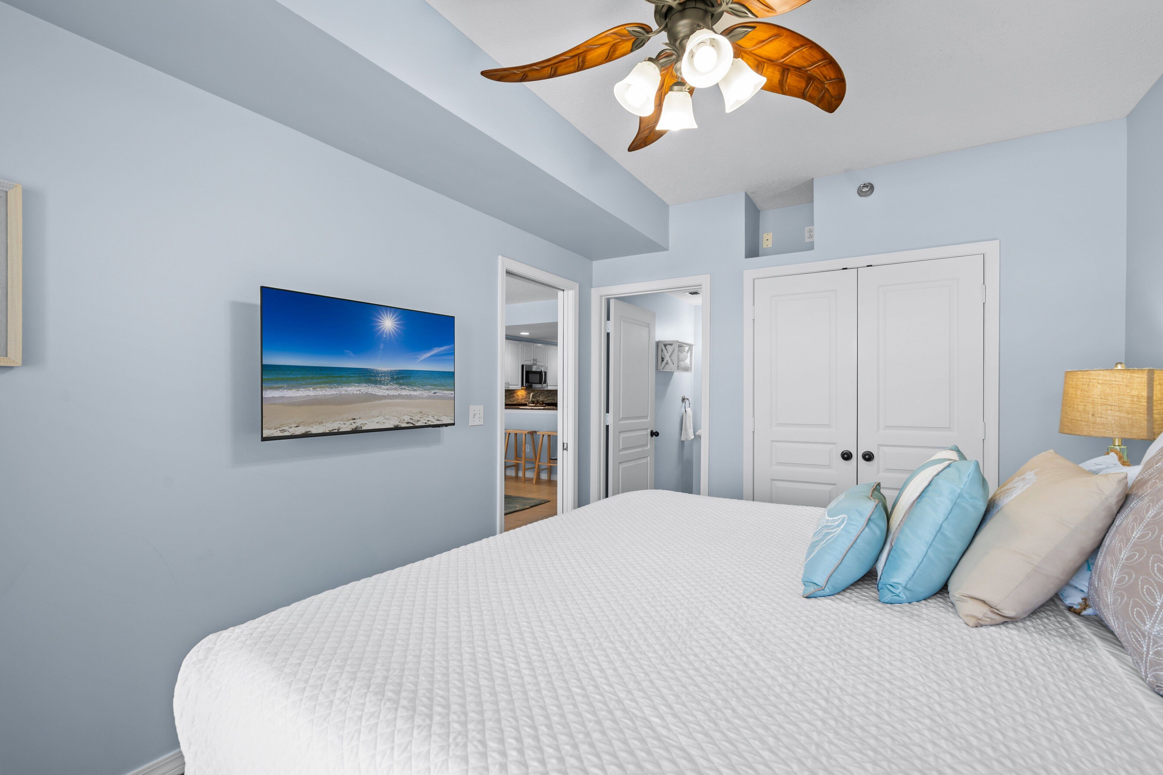 A bedroom with a bed, pillows, a ceiling fan, a wall-mounted TV, and a door leading to a bathroom.
