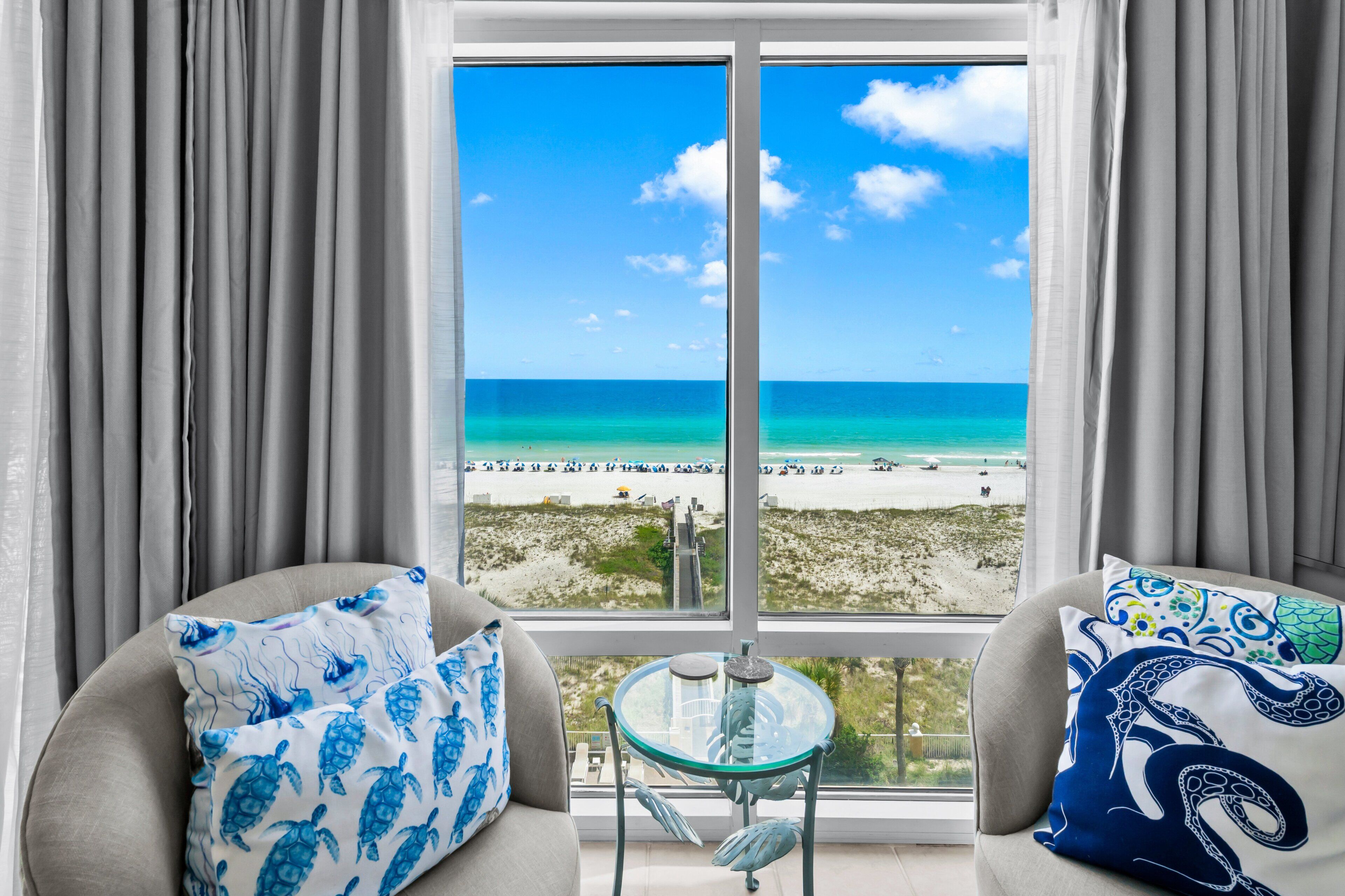 Een kamer met een groot raam dat uitkijkt op een strand, twee banken met gestreepte kussens en een kleine tafel.
