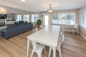 A spacious living area with a large sofa, dining table, and chairs, a kitchen in the background, and a balcony with a view.