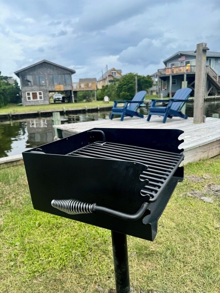 A black barbecue grill with a spring mechanism, set against a backdrop of a residential area with houses and a dock.