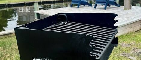 A black barbecue grill with a spring mechanism, set against a backdrop of a residential area with houses and a dock.