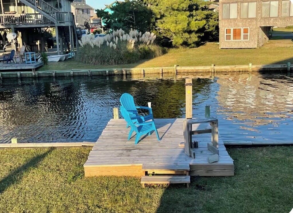A wooden dock with a blue chair and a small table, a grassy area, and a body of water.