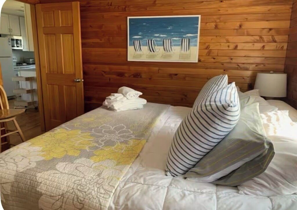 A wooden-paneled room with a bed, a painting of beach chairs, and a door leading to a kitchen.