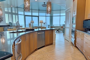 A modern kitchen with a curved island, wooden cabinets, and a view to the outside. - Turquoise Place D2201 | Balcony Hot Tub & Grill (Orange Beach)
