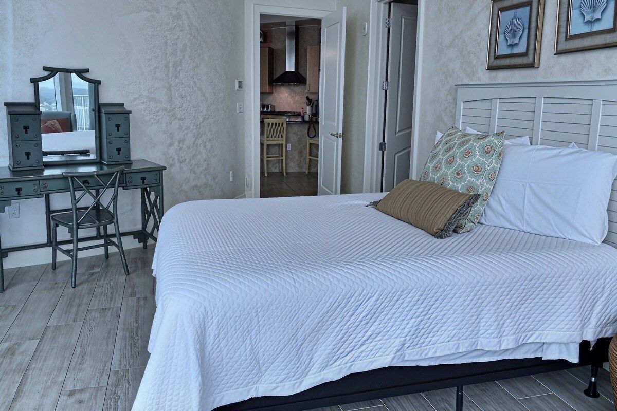 A bedroom with a bed, a desk with a mirror, and a view into the kitchen.
