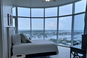 A bedroom with a large bay window, a bed with pillows, a small round table, and a chair. - Turquoise Place D2201 | Balcony Hot Tub & Grill (Orange Beach)