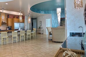 A modern kitchen with a central island, bar stools, and a dining area with wicker furniture. - Turquoise Place D2201 | Balcony Hot Tub & Grill (Orange Beach)