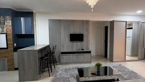 A modern living room with a grey sofa set, a coffee table, a TV mounted on the wall, and a kitchen area with a bar and stools. - Condomínio Henrique I (Balneário piçarras)