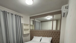 A neatly made bed in a hotel room with a headboard, a bedside table, a mirror, and a wall-mounted air conditioning unit. - Condomínio Henrique I (Balneário piçarras)