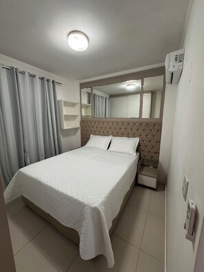 Una cama bien hecha en una habitación de hotel, con cabecera, mesita de noche, espejo y aire acondicionado de pared.