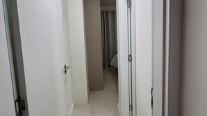 A narrow hallway with white walls and doors, leading to a room with a bed and a window. - Condomínio Henrique I (Balneário piçarras)