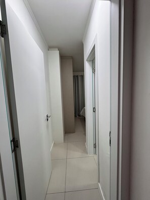 Un pasillo estrecho con paredes y puertas blancas que conduce a una habitación con cama y ventana.