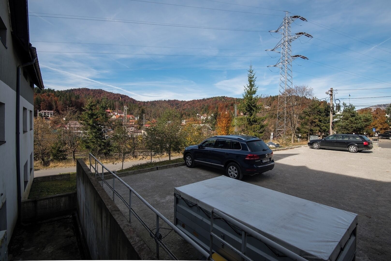 A parking area with a blue car parked in the foreground, a white truck in the background, and a view of a hilly landscape with trees and buildings.