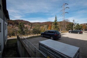 A parking area with a blue car parked in the foreground, a white truck in the background, and a view of a hilly landscape with trees and buildings.