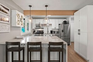 Une cuisine moderne dotée d’un îlot central, de meubles blancs, d’appareils électroménagers en acier inoxydable et de luminaires suspendus.