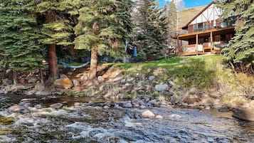 Une cabane dotée d’une terrasse donnant sur un ruisseau, entourée d’arbres.