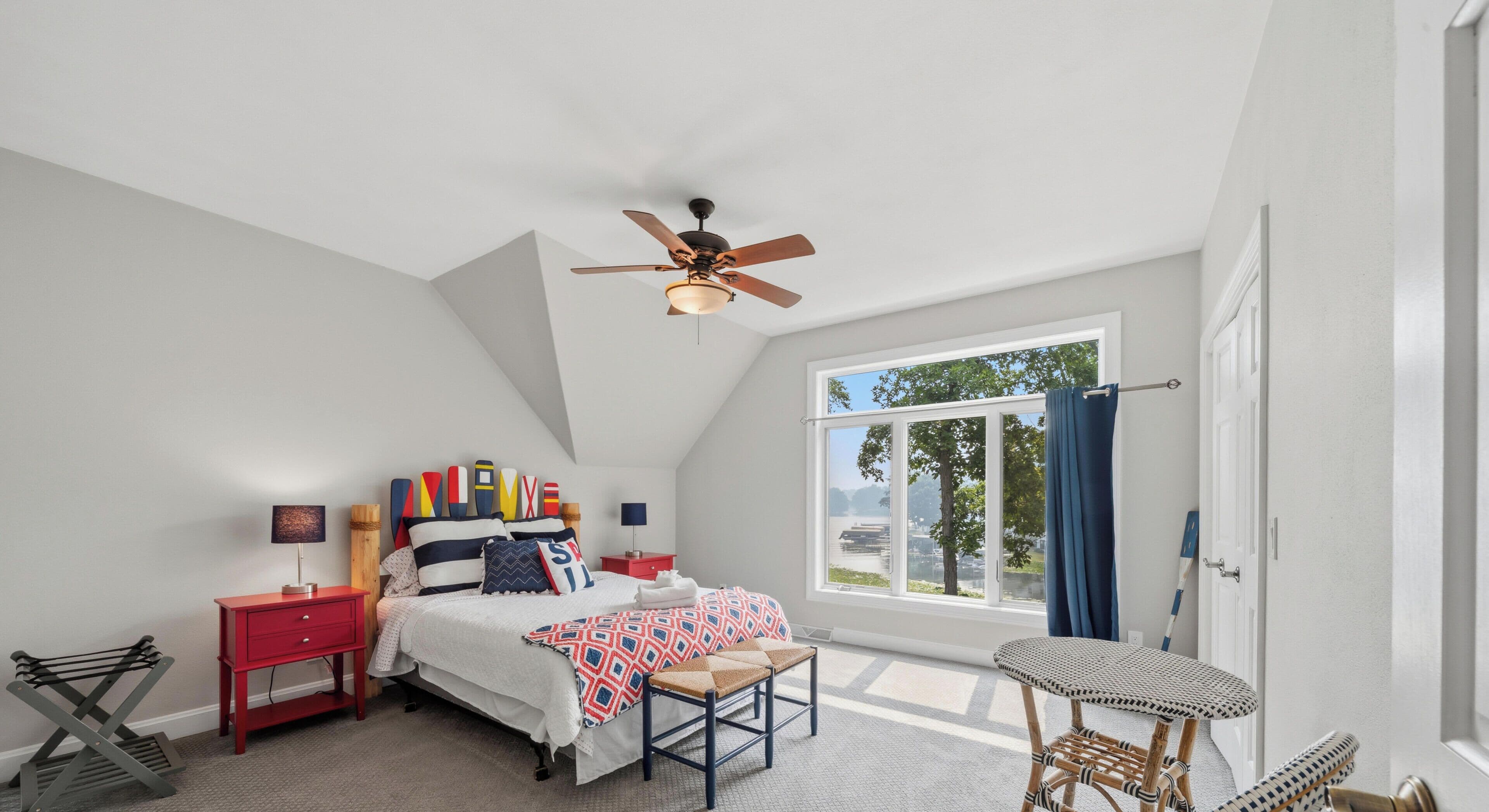 Un dormitorio con cama, mesita de noche, cómoda roja, ventana con vista, ventilador de techo y puerta.