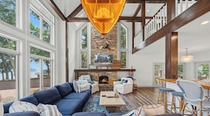 A spacious living room with a fireplace, large windows, and a high ceiling.