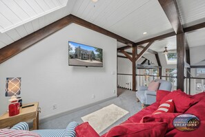 A spacious loft-style room with a high ceiling, exposed wooden beams, a large window, and a view of a house.