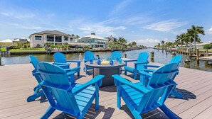 Une terrasse au bord de l’eau, agrémentée de chaises Adirondack bleues, d’une table ronde et offrant une vue dégagée sur une marina, avec des bateaux et des maisons en arrière-plan.