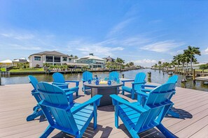 A waterfront deck with blue Adirondack chairs, a round table, and a clear view of a marina with boats and houses in the background. - Modern Waterfront Retreat with Pool, Spa & Canal Access - FMB - Sea-O-Siesta - Roelens (Fort Myers Beach)
