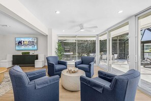A modern living room with a large glass door leading to an outdoor area, a flat-screen TV mounted on the wall, and a ceiling fan. - Modern Waterfront Retreat with Pool, Spa & Canal Access - FMB - Sea-O-Siesta - Roelens (Fort Myers Beach)
