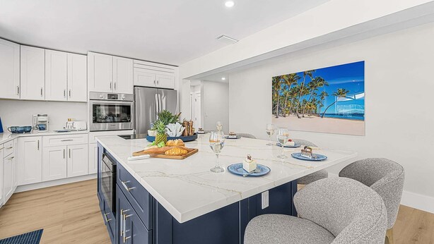 Une cuisine moderne avec des meubles blancs, un grand îlot central doté de chaises bleues et grises, et un tableau représentant une scène de plage.