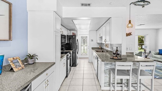 A modern kitchen with white cabinetry, a granite countertop, and a central island with bar stools. - Gulf Access Escape with Heated Pool, Dock & Kayaks - Villa Maria - Roelens (Cape Coral)