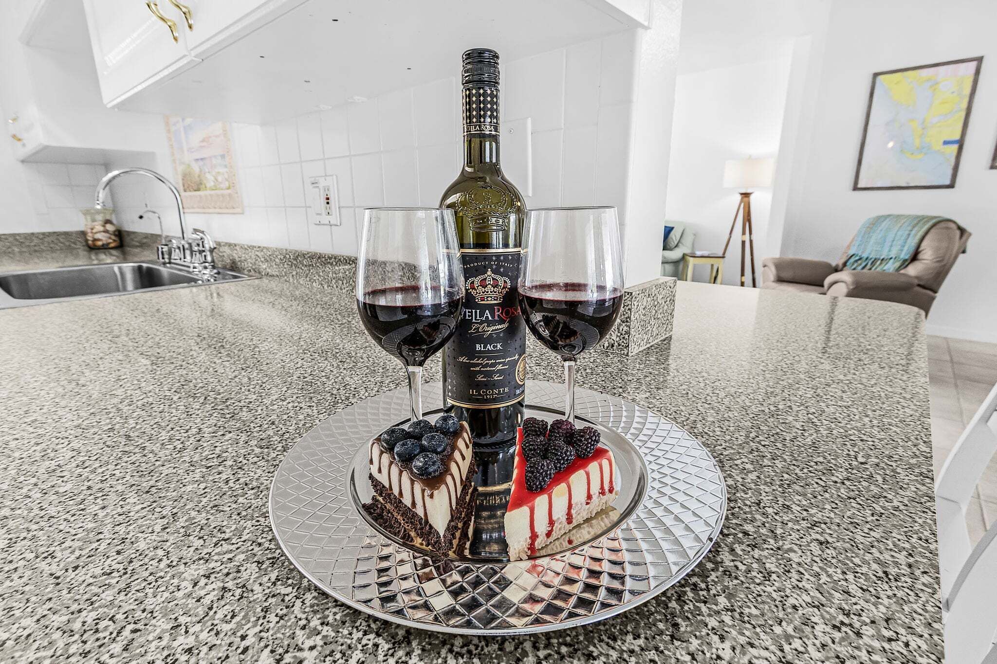 A kitchen counter with a decorative bottle stand holding two glasses of red wine, a cake, and a bottle of Felarosa wine.