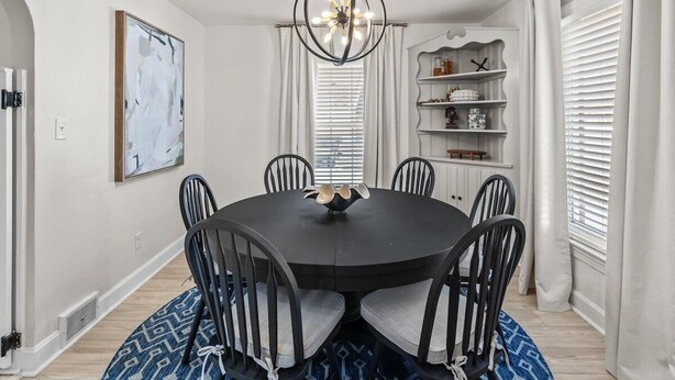 A dining room with a round table, chairs, a chandelier, and a wall-mounted shelf with decorative items.