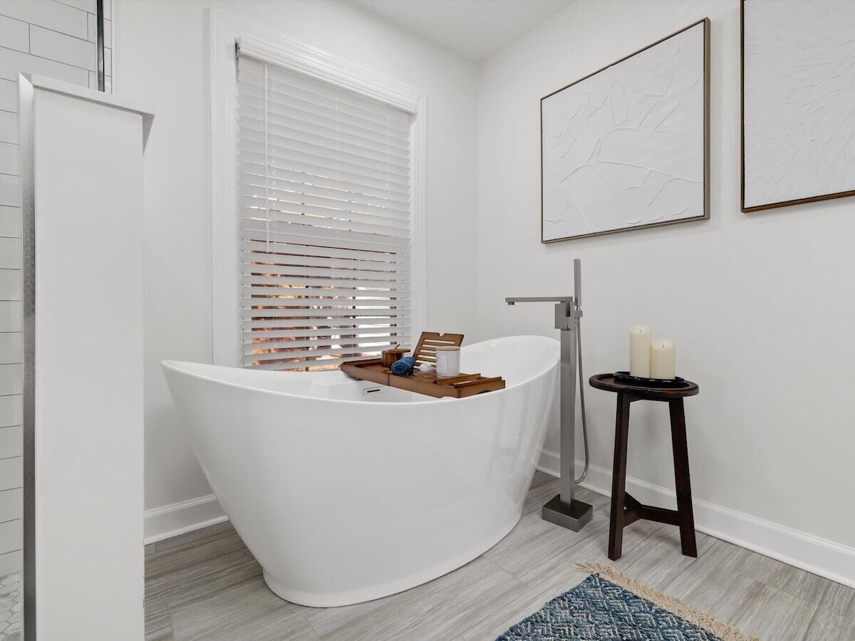 A modern bathroom with a freestanding bathtub, a window with blinds, a small wooden stool with candles, and two framed artworks on the wall.